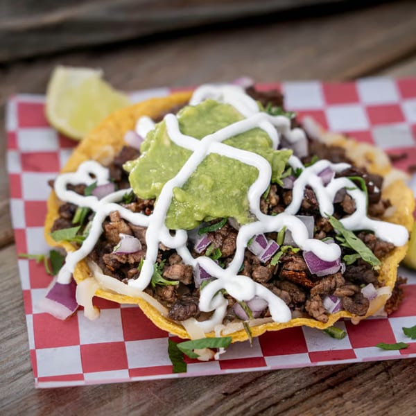 A Tacos El Patron tostada with meat, guacamole, sour cream, and red onions on checkered paper.