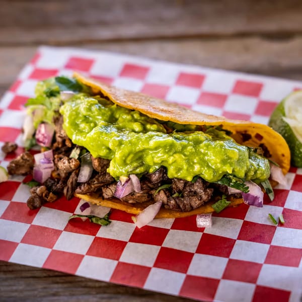A Tacos El Patron carne asada taco with guacamole, onions, and cilantro on checkered paper.