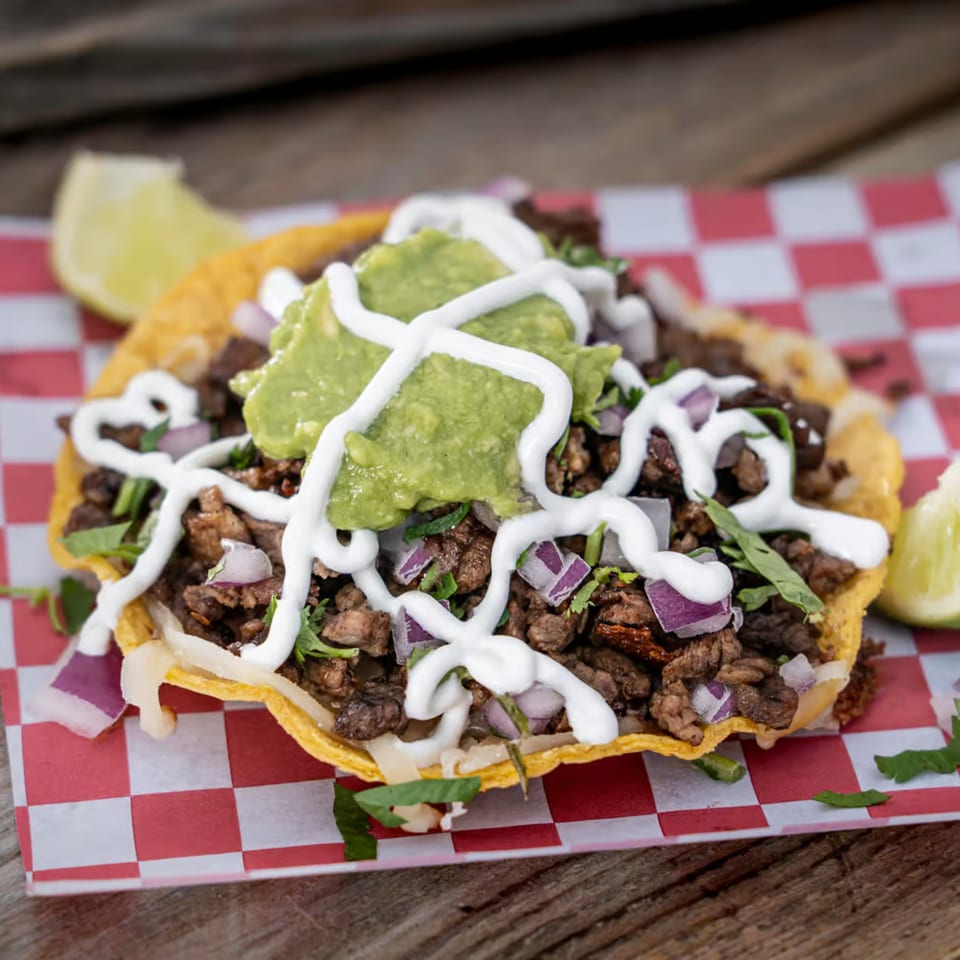 A Tacos El Patron tostada with carne asada, guacamole, sour cream, and red onions.