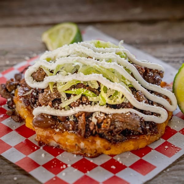 Close-up of a Mexican sopes with meat, lettuce, sour cream, and cheese on checkered paper.
