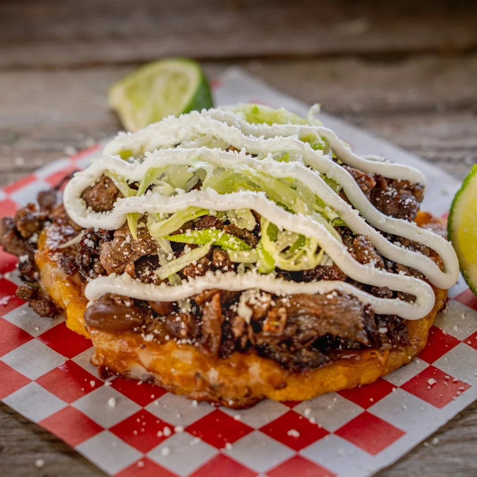 Close-up of a Mexican sopes with meat, lettuce, sour cream, and cheese on checkered paper.