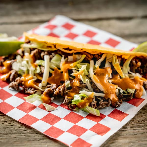 A Tacos El Patron taco with meat, lettuce, and orange sauce on red and white checkered paper.