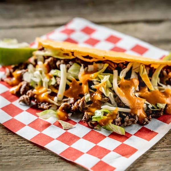 A Tacos El Patron taco with meat, lettuce, and orange sauce on checkered paper.