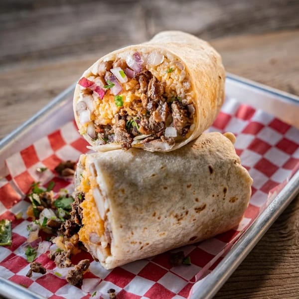 Two halves of a large burrito filled with meat, rice, and vegetables on a checkered paper-lined tray.