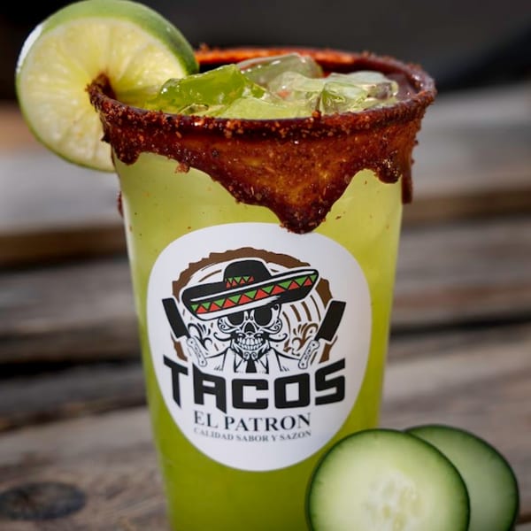A Tacos El Patron branded cup with a green drink, chili-lime rim, lime, and cucumber slices.